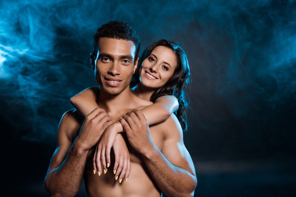 young cheerful woman hugging muscular mixed race man on black with blue smoke